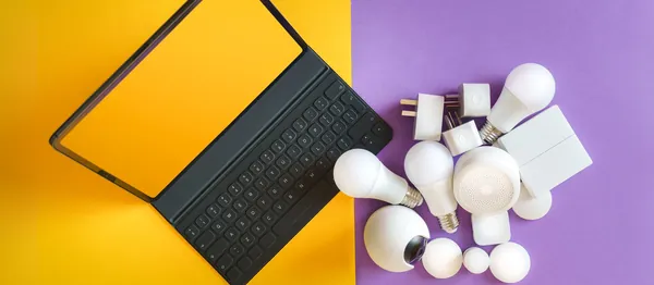 Tablet com teclado sobre fundo amarelo e lilás, ao lado de lâmpadas